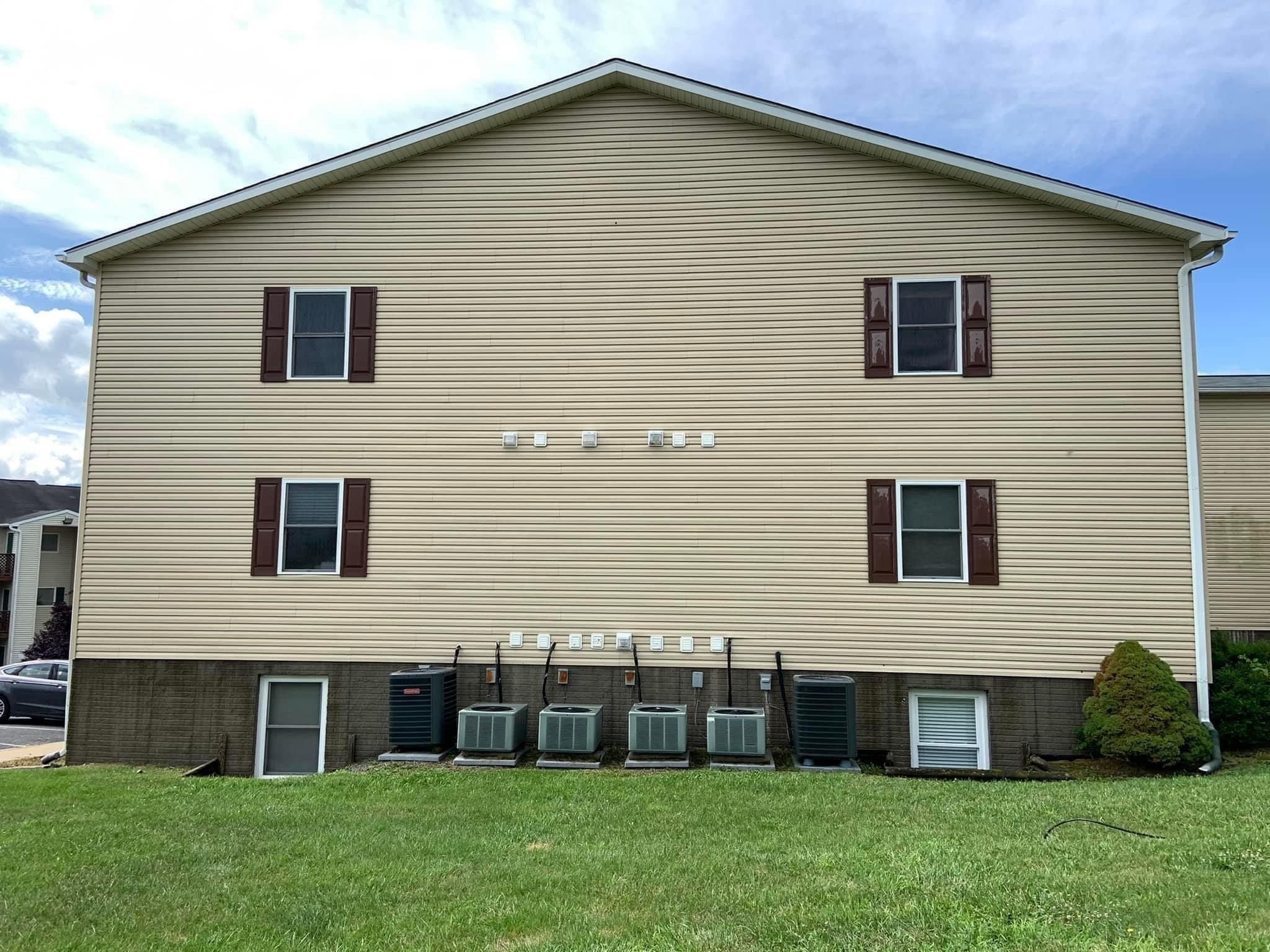 Tan vinyl gable wall of an apartment building after washing, with dark red shutters, small vents, and a row of air conditioning units along the foundation