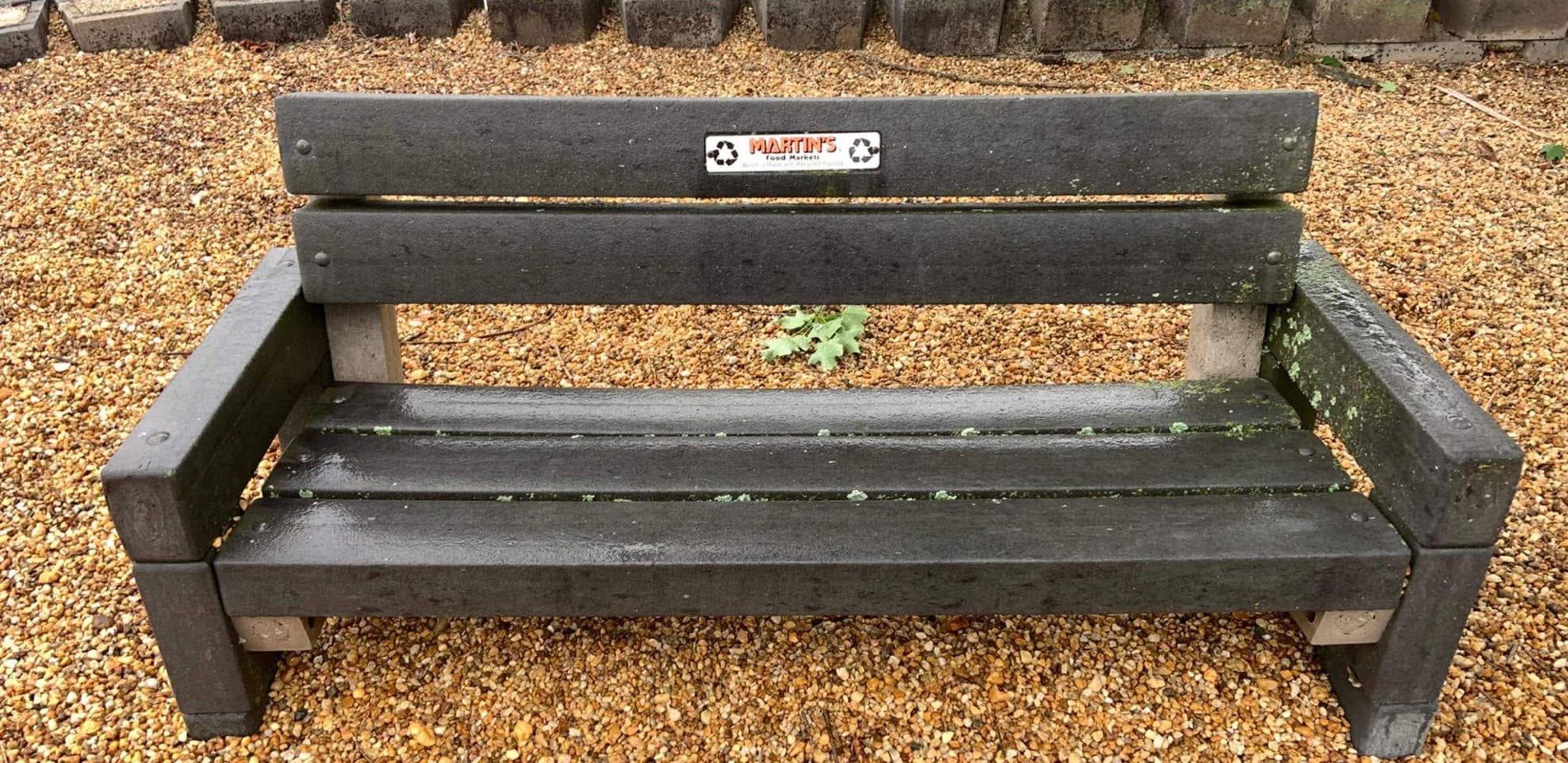 Dark outdoor bench on gravel with green lichen on the arm and seat