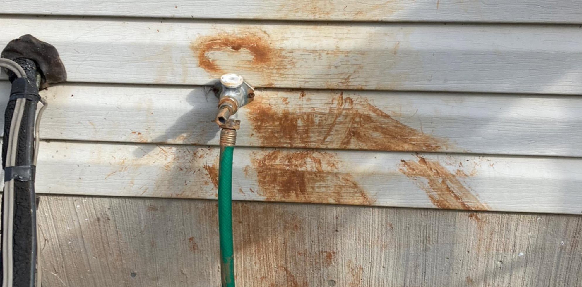 Orange rust stains spreading across white vinyl siding around an outdoor spigot