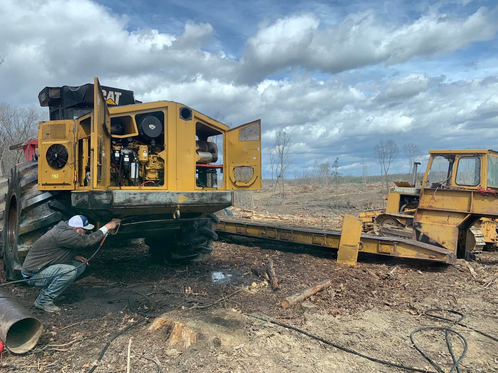 Crew member crouched in a cleared field washing the underside of a yellow logging skidder, its engine doors open
