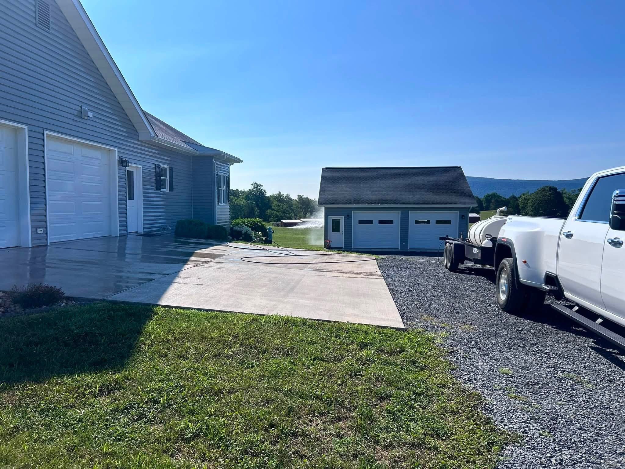 Clean concrete garage apron beside a blue-gray house, a hose looped across the slab, with a white pickup and water tank trailer parked on the gravel and hills behind